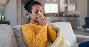 Portrait of young black woman sneezing in to tissue at home. Sick african woman wrapped in blanket sitting on sofa blowing her nose at home. Ill girl sneezing with runny nose in winter.