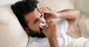 Sick Young Indian Man Blowing Nose In Paper Tissue While Lying In Bed At Home, Ill Eastern Guy Suffering Migraine, Seasonal Flu Or Cold, Having Rhinitis Or Allergic Reaction, Closeup Shot