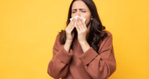 Young sick woman having allergy and sneezing in tissue on yellow background